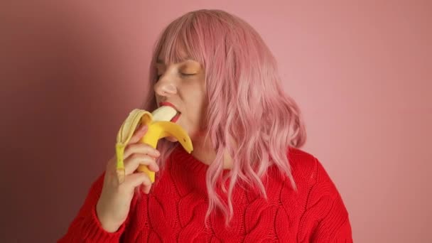 Молодая женщина с розовыми волосами ест банан на розовом фоне. Высококачественные FullHD кадры - Кадры, видео