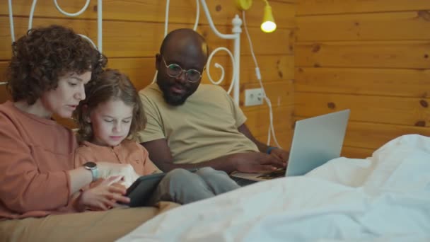 Medium shot van etnisch diverse familie zitten in bed. Blanke moeder helpen haar kind met tablet en vader werken op afstand op laptop naast hen - Video
