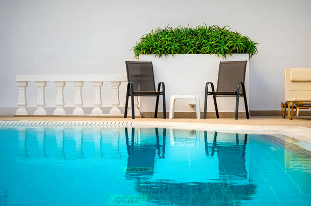 Dos sillas de relajación situadas junto a la piscina de agua cristalina azul. Una pared blanca prístina sirve como telón de fondo adornado con plantas verdes vibrantes, creando un oasis sereno. Relajación definitiva - Foto, Imagen