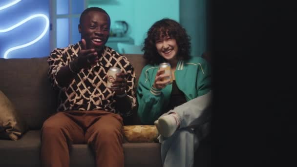 Střední záběr různorodého páru smích a pití limonády, zatímco spolu tráví čas sledováním televize na gauči v neonově osvětleném obývacím pokoji - Záběry, video