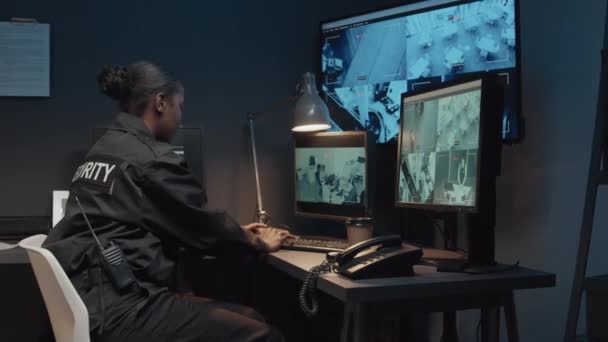 Portret van een jonge lachende Afro-Amerikaanse bewaker in zwart uniform die camerabeelden controleert en dan kijkt naar de camera in de bewakingskamer. - Video