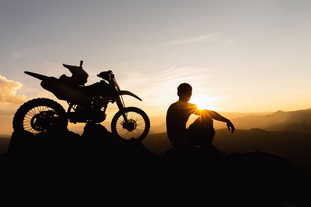 Mann mit Motocross-Rad gegen schöne Lichter, Silhouette eines Mannes mit Motocross-Motorrad Auf dem Gipfel des felsigen Hochgebirges bei schönem Sonnenuntergang, Enduro-Motorrad-Reisekonzept. - Foto, Bild