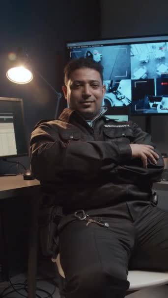 Ritratto verticale di un maschio sorridente Agente di sicurezza biennale seduto nella sala di sorveglianza con riprese video a circuito chiuso sui monitor e che guarda la telecamera - Filmati, video