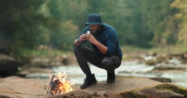 Een reiziger drinkt thee uit een metalen mok bij een kampvuur tijdens een toeristische wandeling in de bergen - Video