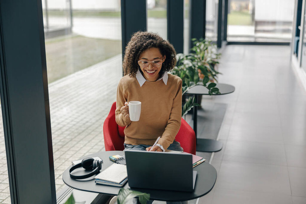 Étudiante souriante travaillant sur un ordinateur portable et prenant des notes tout en buvant du café dans un coworking confortable - Photo, image
