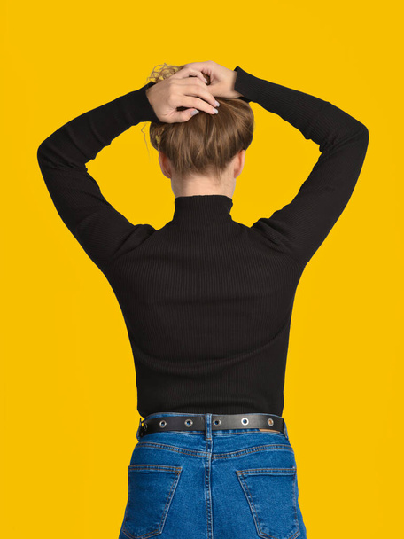 A young Caucasian woman holds her hair collected on her head with her hands. Rear view. Yellow background. - Photo, Image