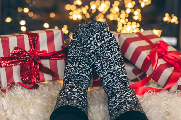 Feet in Christmas socks and gift boxes in front of the fireplace, close up, relaxing at home, cozy background. - Photo, Image