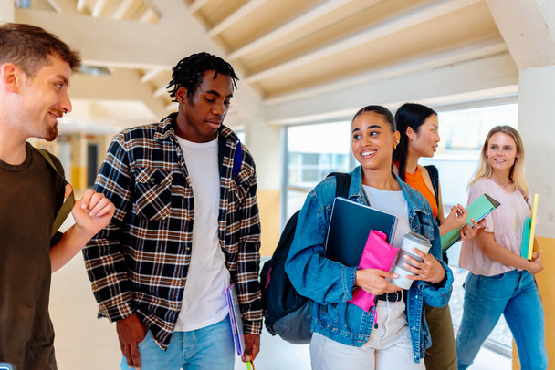 Πολυφυλετική ομάδα μαθητών με έμφαση στη νεαρή Λατίνα κοπέλα που περπατά και συνομιλεί μαζί τους στις αίθουσες της πανεπιστημιούπολης ή του γυμνασίου. συμμαθητές και πίσω στο σχολείο - Φωτογραφία, εικόνα