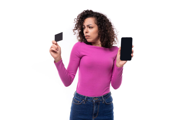 κομψό λεπτή νεαρή σγουρά μελαχρινή γυναίκα ντυμένη με λιλά ζιβάγκο κατέχει μια πιστωτική κάρτα και ένα smartphone. - Φωτογραφία, εικόνα