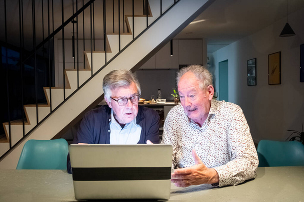 Це зображення захоплює двох літніх чоловіків, глибоко захоплених тим, що, здається, є спільним цифровим досвідом на ноутбуці. Чоловік зліва, з окулярами, нахиляється вперед інтенсивно, можливо, усунення несправностей - Фото, зображення