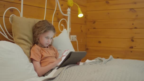 Середній знімок хлопчика з кучерявим волоссям, лежачи в ліжку, прокручуючи стрічку новин або граючи в онлайн гру на цифровому планшеті і граючи в онлайн ігри - Кадри, відео