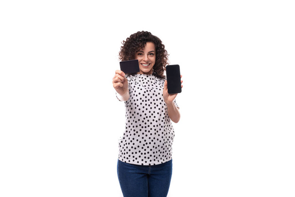 joven mujer caucásica de pelo oscuro vestida con una blusa sostiene una maqueta de tarjeta de plástico y un teléfono inteligente y demuestra a la cámara. - Foto, Imagen