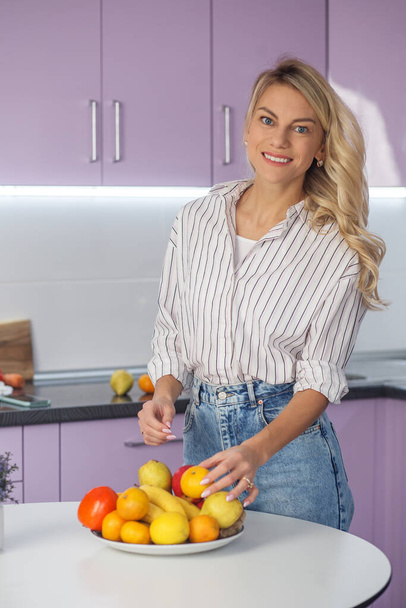 Woman puts fruit on plate in kitchen. Organic food. Millennial woman is preparing food in kitchen. Diet and healthy eating concept. - Photo, Image