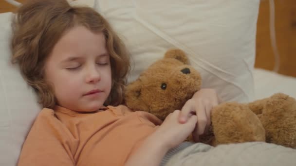 Mediano primer plano de niño con el pelo ondulado que se duerme en su cama con juguete de oso en sus brazos - Metraje, vídeo