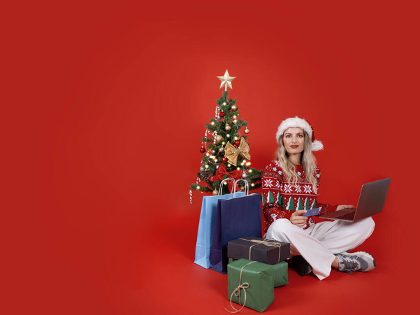 Online-Einkauf zu Weihnachten, fröhliche junge Frau sitzt neben geschmücktem Baum und hält Laptop in der Hand. Verwenden Sie Kreditkarte. Geschenke kaufen, ein frohes neues Jahr Konzeptidee. Roter Hintergrund. - Foto, Bild