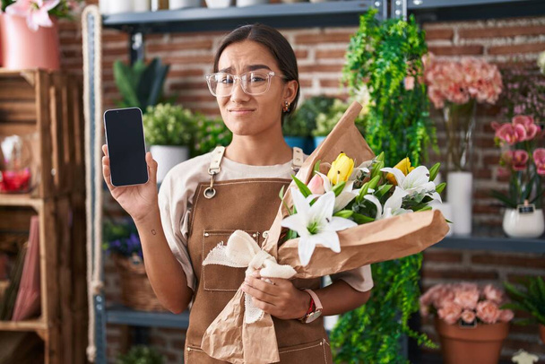 Eine junge hispanische Frau, die in einem Blumenladen arbeitet, zeigt einen ahnungslosen und verwirrten Ausdruck auf dem Smartphone-Bildschirm. Zweifelhaftes Konzept.  - Foto, Bild