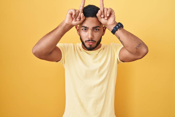 Young hispanic man standing over yellow background doing funny gesture with finger over head as bull horns  - Photo, image