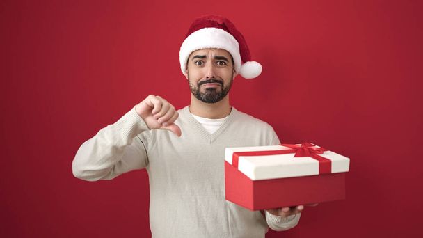 Junger hispanischer Mann mit Weihnachtsmütze hält Geschenk mit gesenktem Daumen über isoliertem roten Hintergrund - Foto, Bild