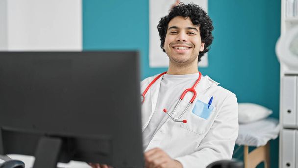 Young latin man doctor using computer smiling at clinic - Photo, Image