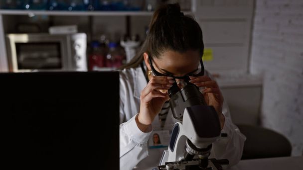 Immersa nella scoperta, bella giovane scienziata ispanica che lavora fino a tarda notte in laboratorio, immersa nella ricerca medica, sperimentando con provette sotto l'analisi ingrandita del microscopio - Foto, immagini