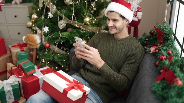 Young hispanic man using smartphone sitting on sofa by christmas tree at home - Foto, Imagem