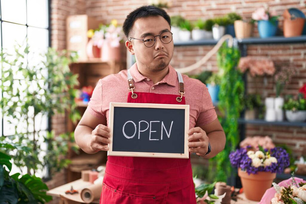 Chinese young man working at florist holding open sign relaxed with serious expression on face. simple and natural looking at the camera.  - Photo, Image