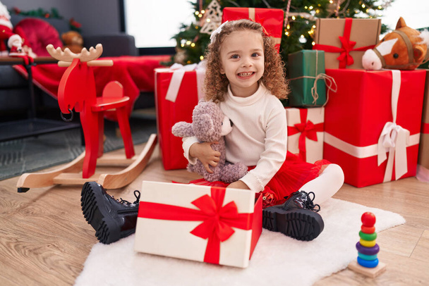 Adorable fille blonde déballer cadeau ours en peluche assis sur le sol par arbre de Noël à la maison - Photo, image