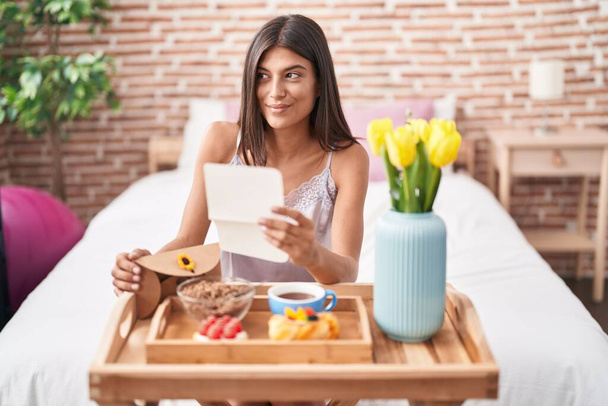 Brunette young woman eating breakfast in the bed reading a letter smiling looking to the side and staring away thinking.  - Photo, Image