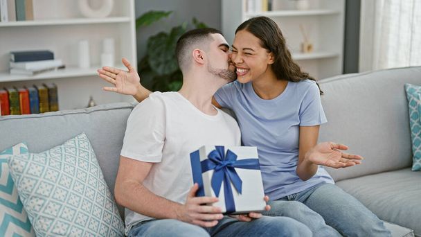 Bella coppia gode bella sorpresa, baciare sul divano, seduti insieme a casa; regalo perfetto approfondisce espressione d'amore in ambiente soggiorno - Foto, immagini