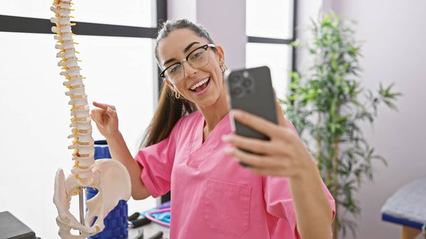 Femme hispanique souriante, physiothérapeute pointant vers le modèle de colonne anatomique lors d'un appel vidéo à son lieu de travail clinique de réadaptation - Photo, image