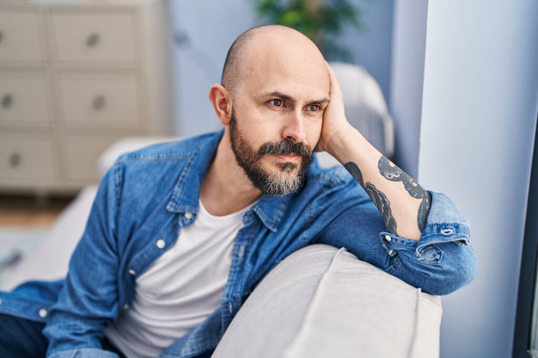 Young bald man sitting on sofa with serious expression at home - Photo, Image