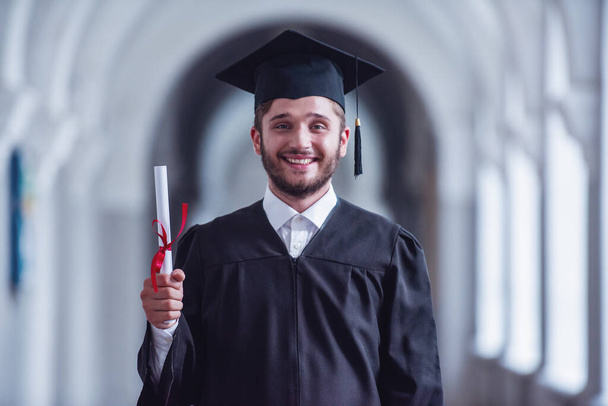 Bonito jovem graduado em vestido acadêmico está segurando diploma, olhando para a câmera e sorrindo enquanto está de pé na sala da universidade
 - Foto, Imagem