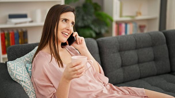 Genç, güzel, İspanyol bir kadın akıllı telefonda konuşuyor. Evde kahve içiyor. - Fotoğraf, Görsel