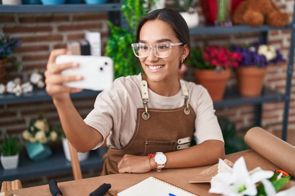 Young beautiful hispanic woman florist make selfie by smartphone sitting on table at florist - Foto, imagen