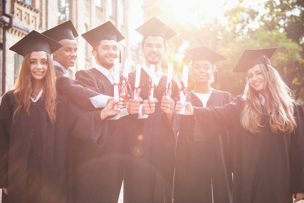 Успешные выпускники в академических платьях имеют дипломы, смотрят в камеру и улыбаются стоя на улице.
 - Фото, изображение