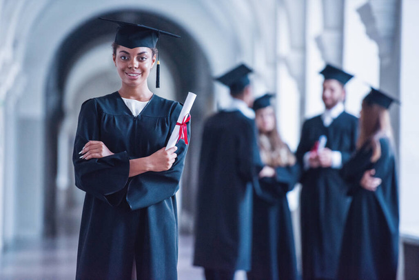 Graduados bem sucedidos em vestidos acadêmicos estão falando na sala da universidade, menina em primeiro plano está segurando diploma, olhando para a câmera e sorrindo
 - Foto, Imagem