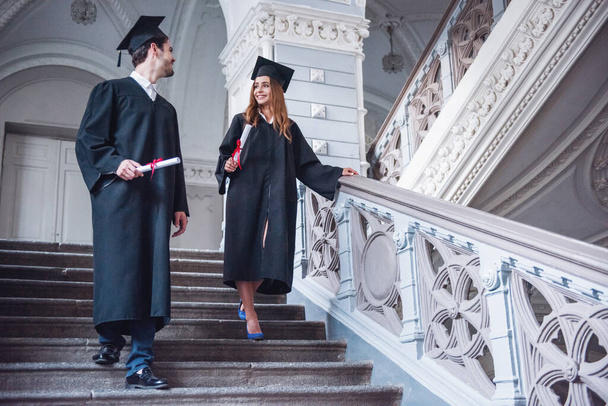 Exitosa pareja de graduados en vestidos académicos están sosteniendo diplomas, hablando y sonriendo mientras van abajo en el edificio de la universidad
 - Foto, imagen