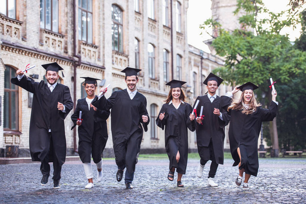Graduados bem sucedidos em vestidos acadêmicos estão segurando diplomas, olhando para a câmera e sorrindo ao correr ao ar livre
 - Foto, Imagem