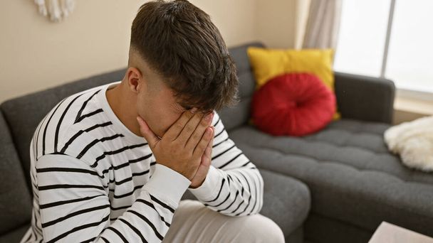 Przygnębiony, zestresowany młody latynoski mężczyzna siedzący samotnie, karmiący ból głowy na domowej sofie, rozpacz nadciągająca w tle jego mieszkania. - Zdjęcie, obraz