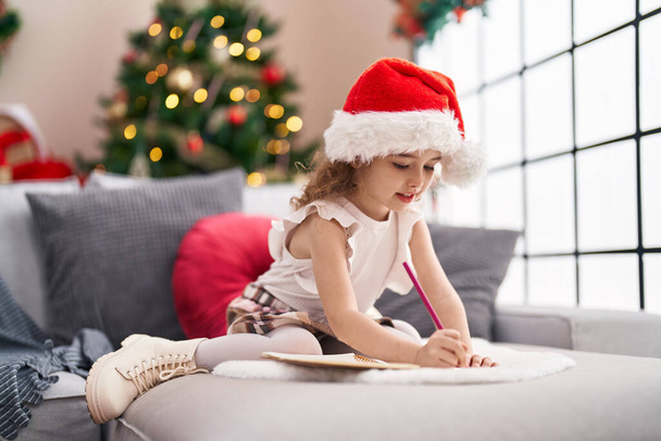 Adorable caucasian girl drawing on notebook sitting on sofa by christmas tree at home - Photo, Image