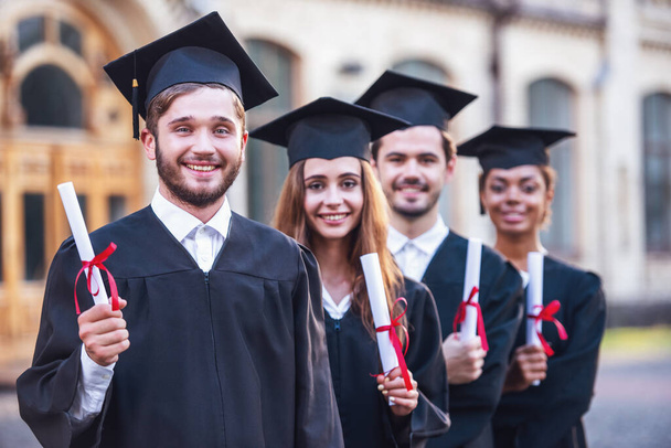 erfolgreiche Absolventen in akademischen Kleidern halten Diplome in der Hand, blicken in die Kamera und lächeln im Freien - Foto, Bild