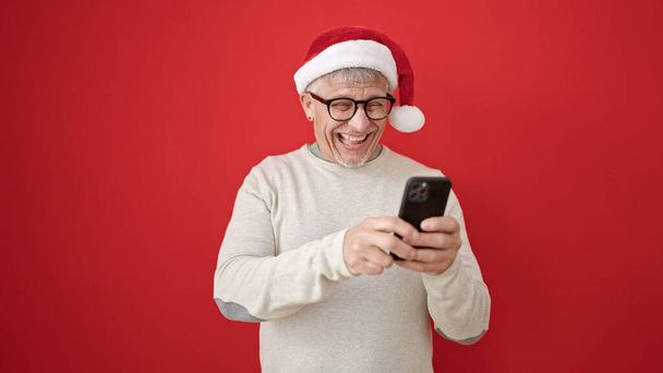 Μεσήλικας γκριζομάλλης άντρας που φοράει χριστουγεννιάτικο καπέλο χρησιμοποιώντας smartphone σε απομονωμένο κόκκινο φόντο - Φωτογραφία, εικόνα