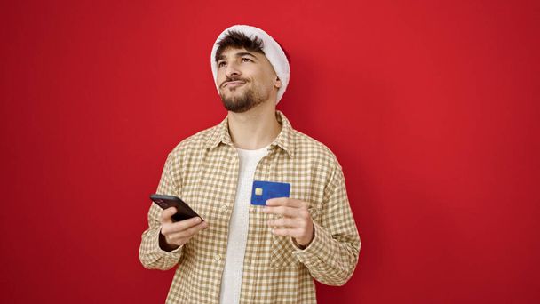 Fiatal arab férfi csinál karácsony online vásárlás okostelefon felett elszigetelt piros háttér - Fotó, kép