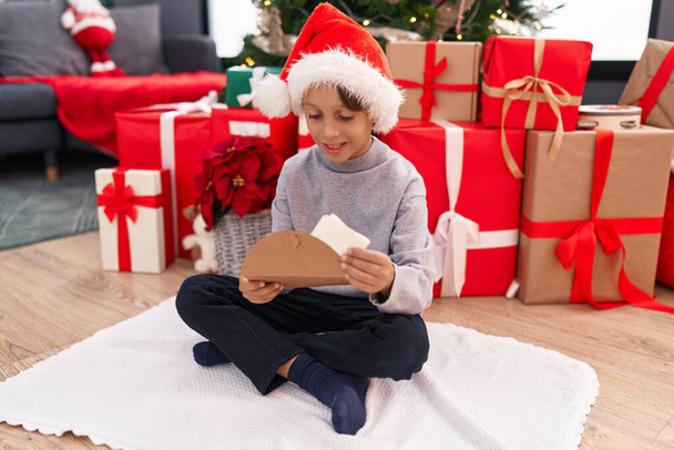 Adorable garçon hispanique mettant lettre Santa Claus dans l'enveloppe assis par arbre de Noël à la maison - Photo, image