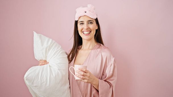 単離されたピンクの背景上のコーヒーと枕を保持する睡眠マスクを着用している若い美しいヒスパニックの女性 - 写真・画像