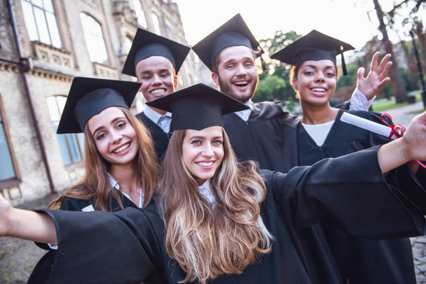 Succesvol afgestudeerden in academische jurken zijn bezit van diploma's, camera kijken en glimlachen terwijl permanent buitenshuis - Foto, afbeelding