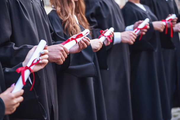 Les diplômés qui réussissent dans les robes universitaires sont titulaires de diplômes, impatients et souriants tout en se tenant debout dans une rangée à l'extérieur
 - Photo, image