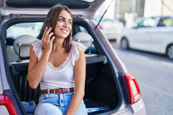 Jovem bela mulher hispânica falando no smartphone sentado no porta-malas do carro na rua - Foto, Imagem