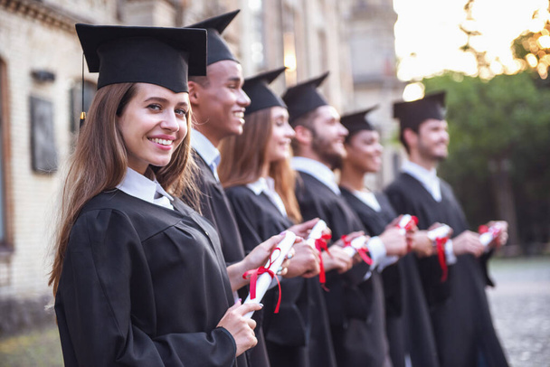 Graduados de sucesso em vestidos acadêmicos estão segurando diplomas, olhando para a frente e sorrindo enquanto está em uma fileira ao ar livre
 - Foto, Imagem