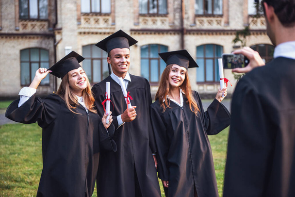 Успешные выпускники в академических платьях держат дипломы, смотрят в камеру и улыбаются стоя на улице, парень фотографирует их
 - Фото, изображение